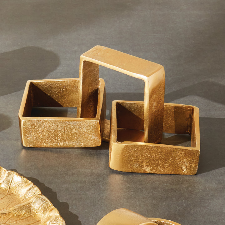Gold chain-link sculpture on a charcoal tabletop, with three square aluminum links beside gold accessories - Modern Organic