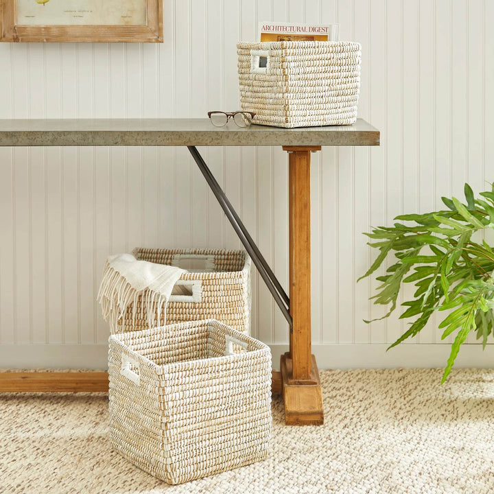 Square woven storage baskets with handles styled under table with plant, used for organizing living room or entryway spaces.


