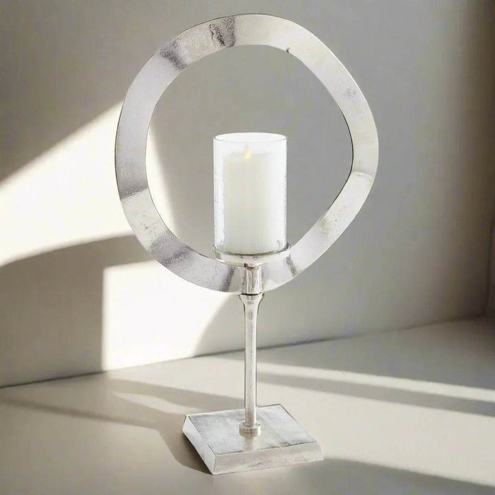 Tall silver candle stand with glass hurricane and LED candle styled in sunlight on tabletop, modern sculptural decor for home – Minimalist Glam.

