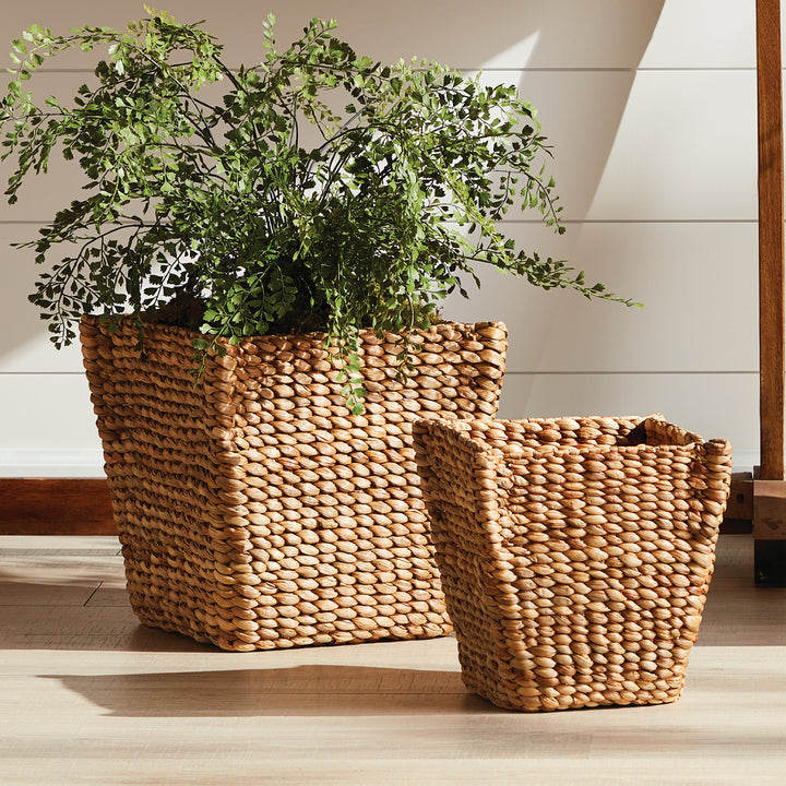 Tamsin tapered basket pair on a floor with leafy stems in the larger basket beside wood furniture and white paneling. Coastal
