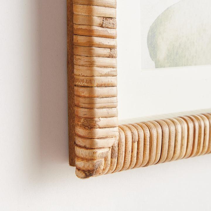 Close view of a natural rattan-wrapped frame corner around a green botanical print, showing its woven textured edge. Coastal