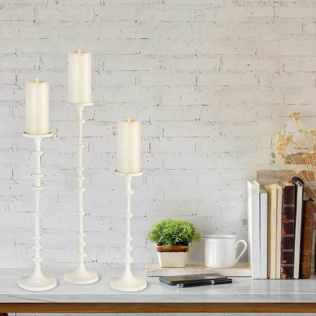 Tall white pillar candle stands on a shelf against a white brick wall with books and greenery, graduated heights add vertical interest – Modern Farmhouse.