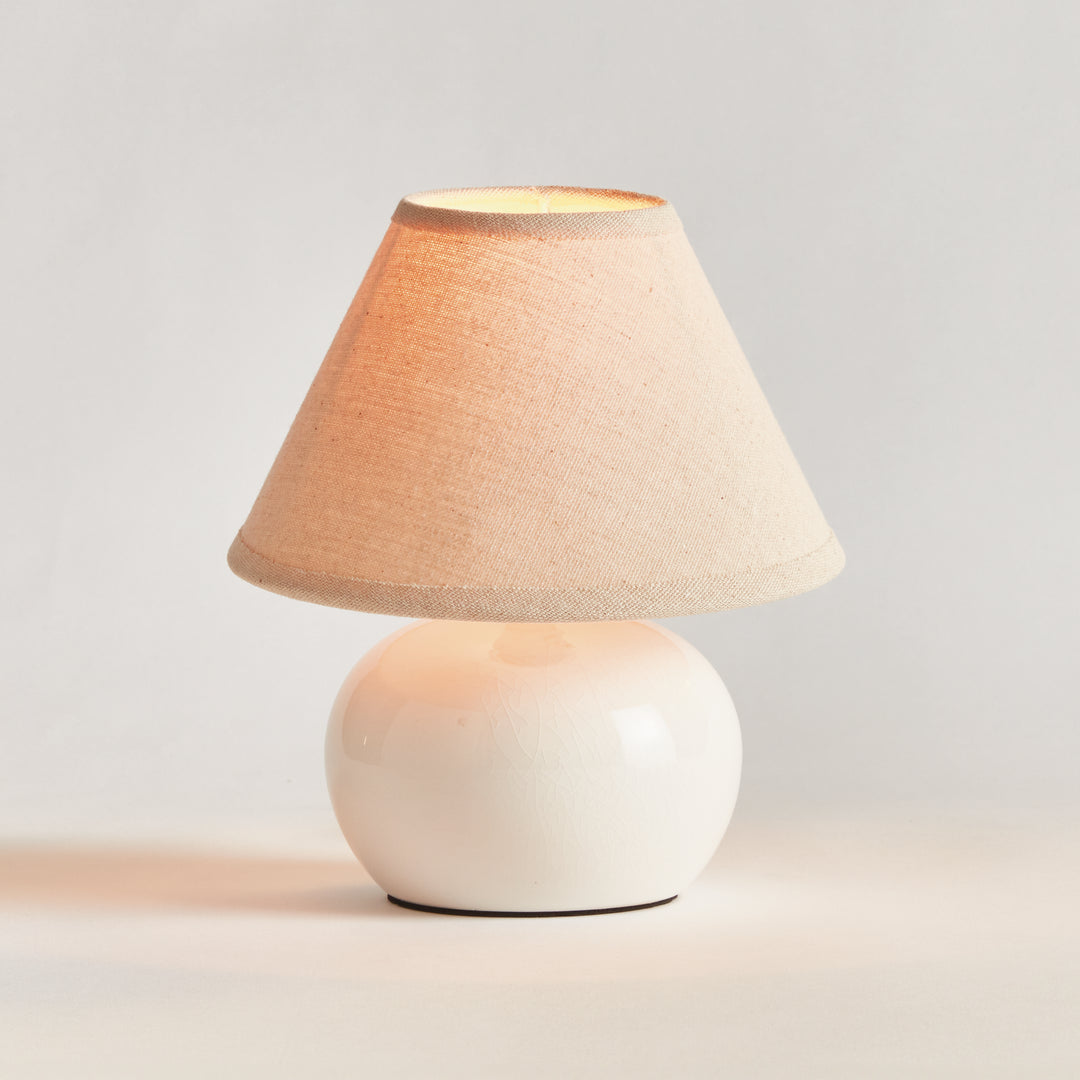 White ceramic mini table lamp illuminated beneath its cream linen shade, shown on a plain backdrop - Coastal Transitional