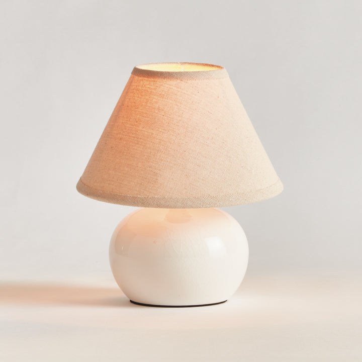 White ceramic mini table lamp illuminated beneath its cream linen shade, shown on a plain backdrop - Coastal Transitional