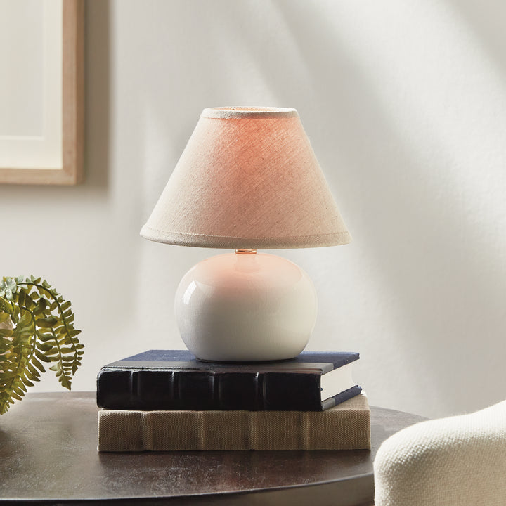 White ceramic mini table lamp lit on stacked books beside a fern, with a glowing cream linen shade - Coastal Transitional