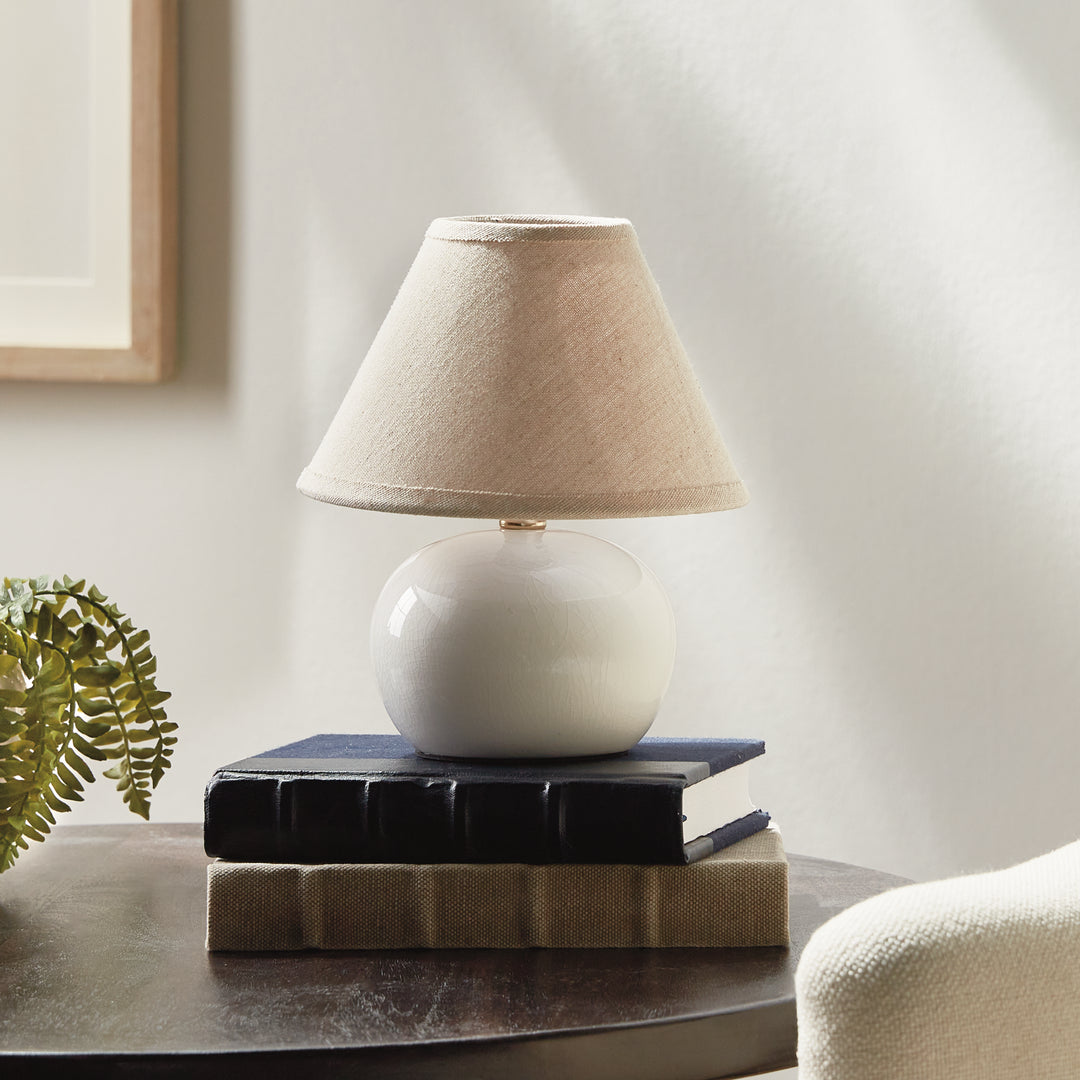 White ceramic mini table lamp on stacked books beside a fern, with a rounded base and linen lampshade - Coastal Transitional