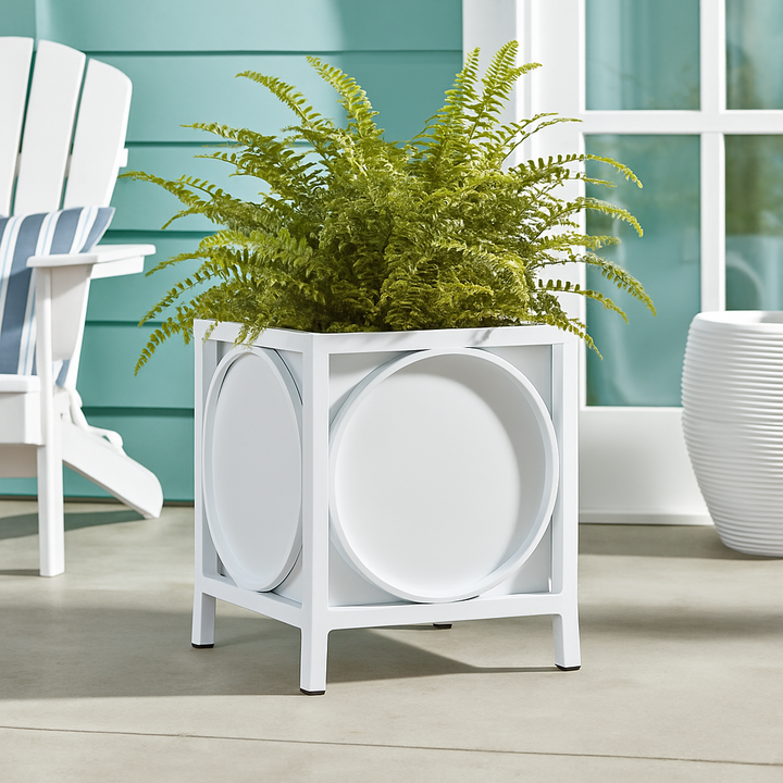 White metal planter with green fern styled on a sunlit coastal terrace beside a rattan chair and blue paneled wall.