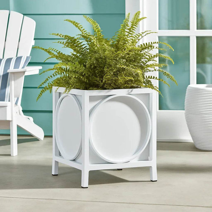 White metal planter with green fern styled on a sunlit coastal terrace beside a rattan chair and blue paneled wall.