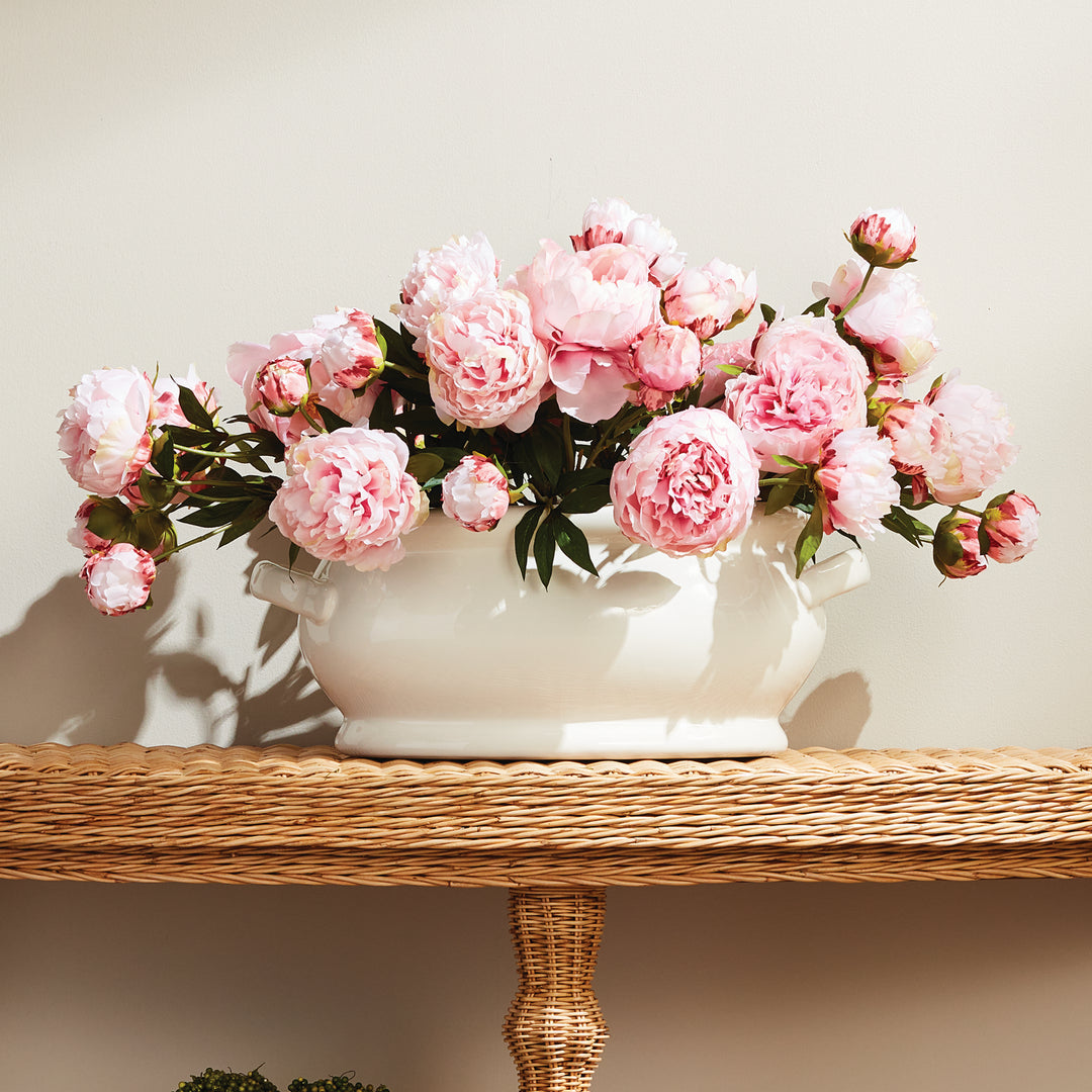 White oval ceramic cachepot filled with pink peonies on a woven console, with sunlight across its curved sides - Traditional