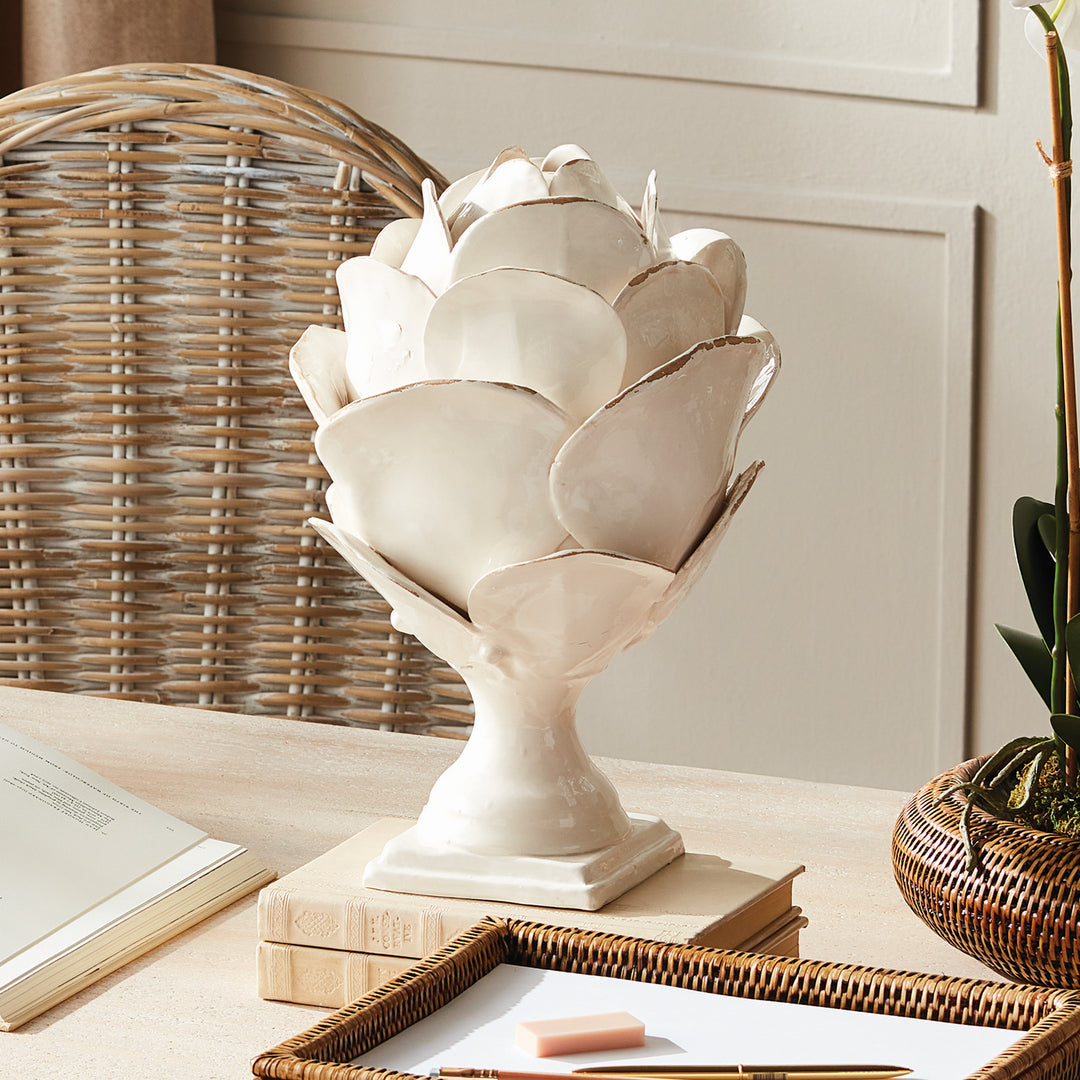 White ceramic petal finial on a desk beside books and a woven chair, showing its layered form and pedestal base, Traditional