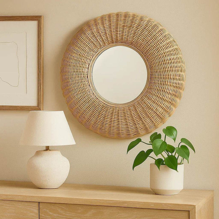 Whitewashed round rattan wall mirror styled above a light wood console with a lamp, framed art, and heartleaf plant in soft lighting.