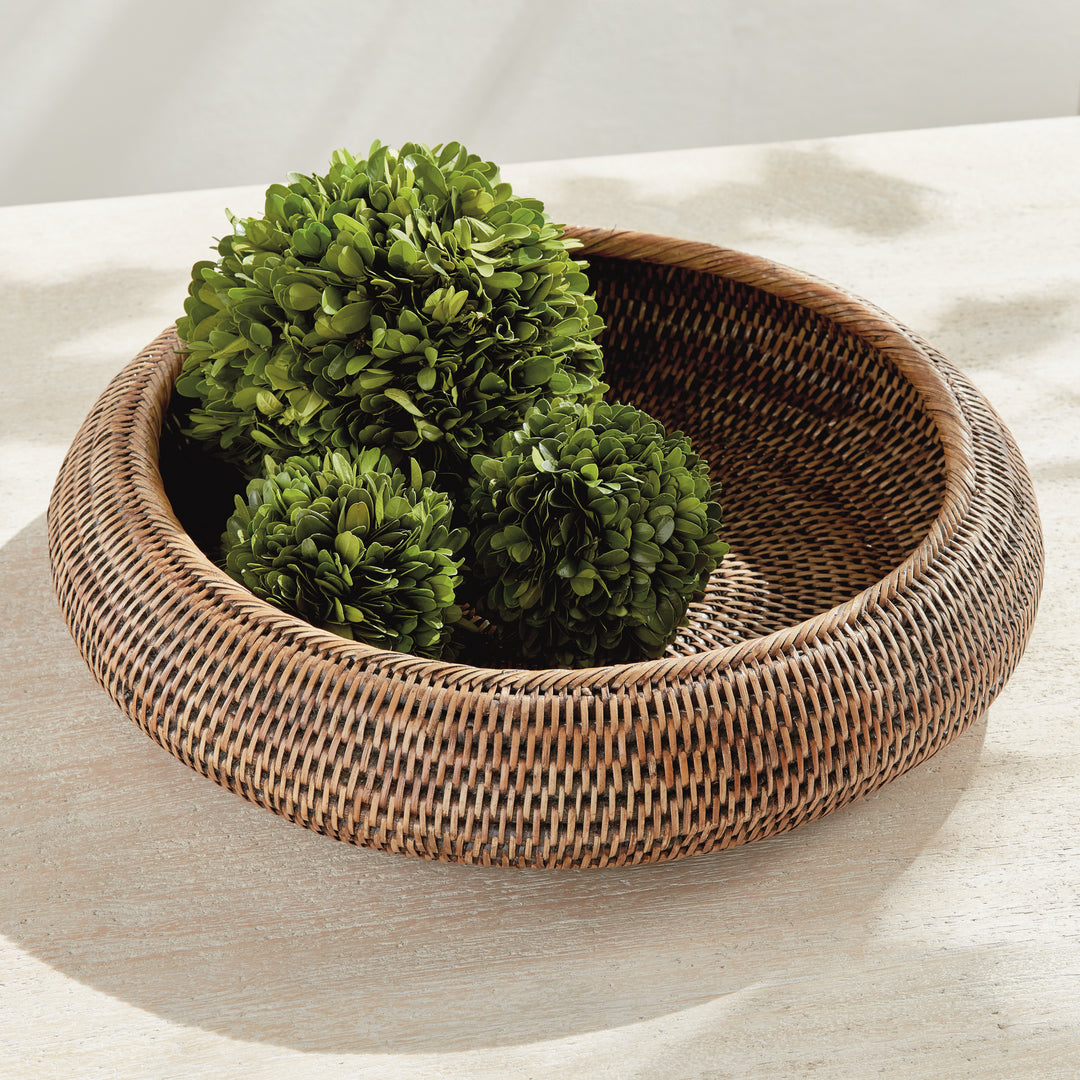 Brown rattan shallow display bowl holding three leafy green decorative balls on a pale stone tabletop - Coastal Transitional