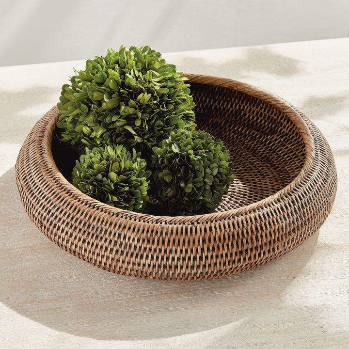 Brown rattan shallow display bowl holding three leafy green decorative balls on a pale stone tabletop - Coastal Transitional